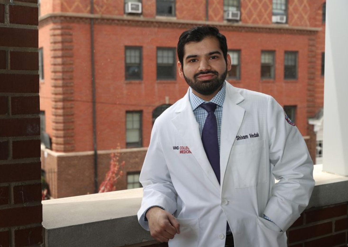 A UIC medical graduate stands by a window, he is wearing a white doctors coat embroidered with the UIC Med school logo on the right breast side of his coat 